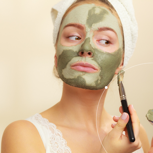 Woman applying a green facial mask with a brush on a beige background