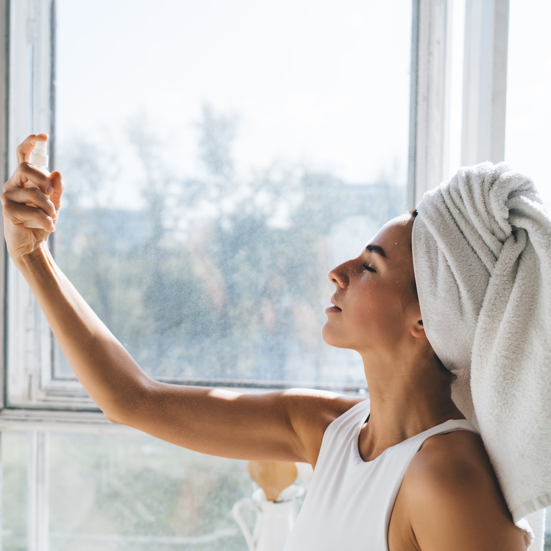 Woman with a towel wrapped around her head, standing by a window with a view of trees. Using facial mist